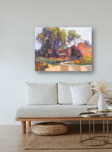 Impressionistic painting depicting a sunlit adobe building with a cross, surrounded by lush greenery and a large tree, mounted on a light wall above a neutral sofa.