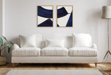 Two framed canvas art pieces with abstract navy blue shapes on white backgrounds hang side-by-side above a cream three-seat sofa in a minimalist living room.