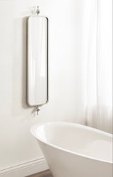 Tall, narrow rectangular mirror with rounded corners and a thin metal frame mounted on a white wall above a white bathtub in a bright bathroom.