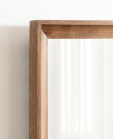 Close-up of a smooth, medium-toned wooden frame corner with clean mitered edges surrounding a reflective mirror surface against a light wall.