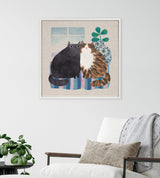 Framed canvas art of two stylized cats sitting side by side on a striped cushion, with a leafy potted plant and window behind them, displayed above a white armchair.