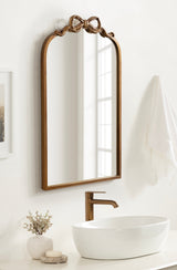 Arched wall mirror with a wooden frame featuring a carved ribbon bow at the top, reflecting a window and placed above a white oval sink and bronze faucet.
