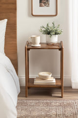 Compact wood side table with curved edges, featuring a potted plant and cream mug on top, a bowl and stacked books on lower shelf, set beside a bed.