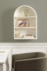 White arched wall shelf with three levels holding a small vase with flowers, a framed family photo, and books with reading glasses, mounted on a gray wall above a dark gray chair.