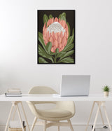 Framed canvas featuring a stylized pink protea flower with white beaded center and green leaves on a black background, hanging above a white desk setup.