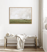 Framed landscape painting with soft green fields and a cloudy sky above, hung on a white wall above a wooden bench with a light fabric draped over it.