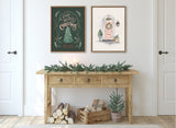 Two framed artworks above a rustic wooden console with pine garland; left art shows green chalkboard-style Christmas tree and text, right art depicts a pink door with holiday wreath.