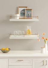 Two white floating shelves with brass railing hold framed photos, glassware, and a pitcher with orange slices, above a white countertop with fruit bowl and flowers.