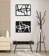 Two framed abstract canvas art pieces with bold black and white strokes hung vertically on a beige wall, above a dark cushioned bench and next to a modern coat rack.