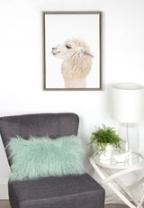 Profile view of a white alpaca with fluffy fur in a simple dark frame hanging above a gray chair with a light green fuzzy pillow and a white side table with a small plant and white lamp.