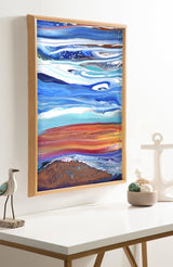 Framed abstract art with flowing horizontal bands of blue, white, and rust red hues resembling a coastal landscape, hanging above a white table with nautical decor.