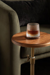 Clear textured glass tumbler with amber liquid on round wooden side table with brass adjustable support next to olive green upholstered sofa