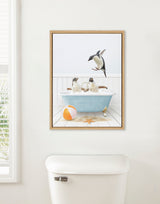 Framed canvas showing three penguins in and around a blue clawfoot bathtub with bubbles, a beach ball, and a starfish on a white wooden floor.