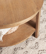 Close-up of a round wooden coffee table with a smooth top and lower shelf, showing natural wood grain and medium brown finish against a cream patterned rug.