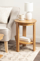 Round wooden side table with four straight legs and a lower shelf holding books, topped with a white textured lamp and a cream coffee mug, next to a light gray sofa.