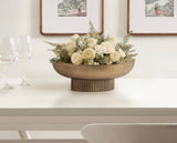 Round bronze-toned ribbed metal bowl centerpiece on white table holding a floral arrangement of white roses and greenery, set near two empty wine glasses.