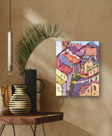 Colorful abstract village painting with geometric houses in purple, red, yellow, and blue tones displayed on a beige wall above a wooden table with vases and a palm leaf.