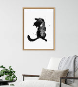 Framed black and white watercolor-style print of a playful cat with splattered ink effects, hanging above a white cushioned chair with a beige pillow and gray throw.
