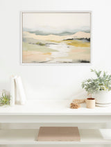 Muted abstract landscape painting with soft blue, beige, and green tones in a white frame hanging above a white shelf with potted plants and books.