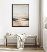 Framed abstract landscape painting with soft beige, gray, and light blue tones hanging above a wooden bench with an open book and draped white fabric in a bright room.