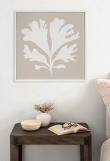 Framed coral beige canvas with a large white abstract leaf design hangs above a dark wooden side table holding a white vase with twigs, a small bowl, and an open book with glasses.