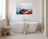 Framed canvas of a small red rowboat floating on calm blue water, hung above a freestanding white bathtub with a beige towel draped over the side in a minimal light-toned bathroom.