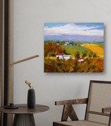 Canvas print of a vibrant rural landscape with green and golden fields, white houses with red roofs, and distant blue mountains under a cloudy sky, hung on a gray wall above a wooden chair and side table.
