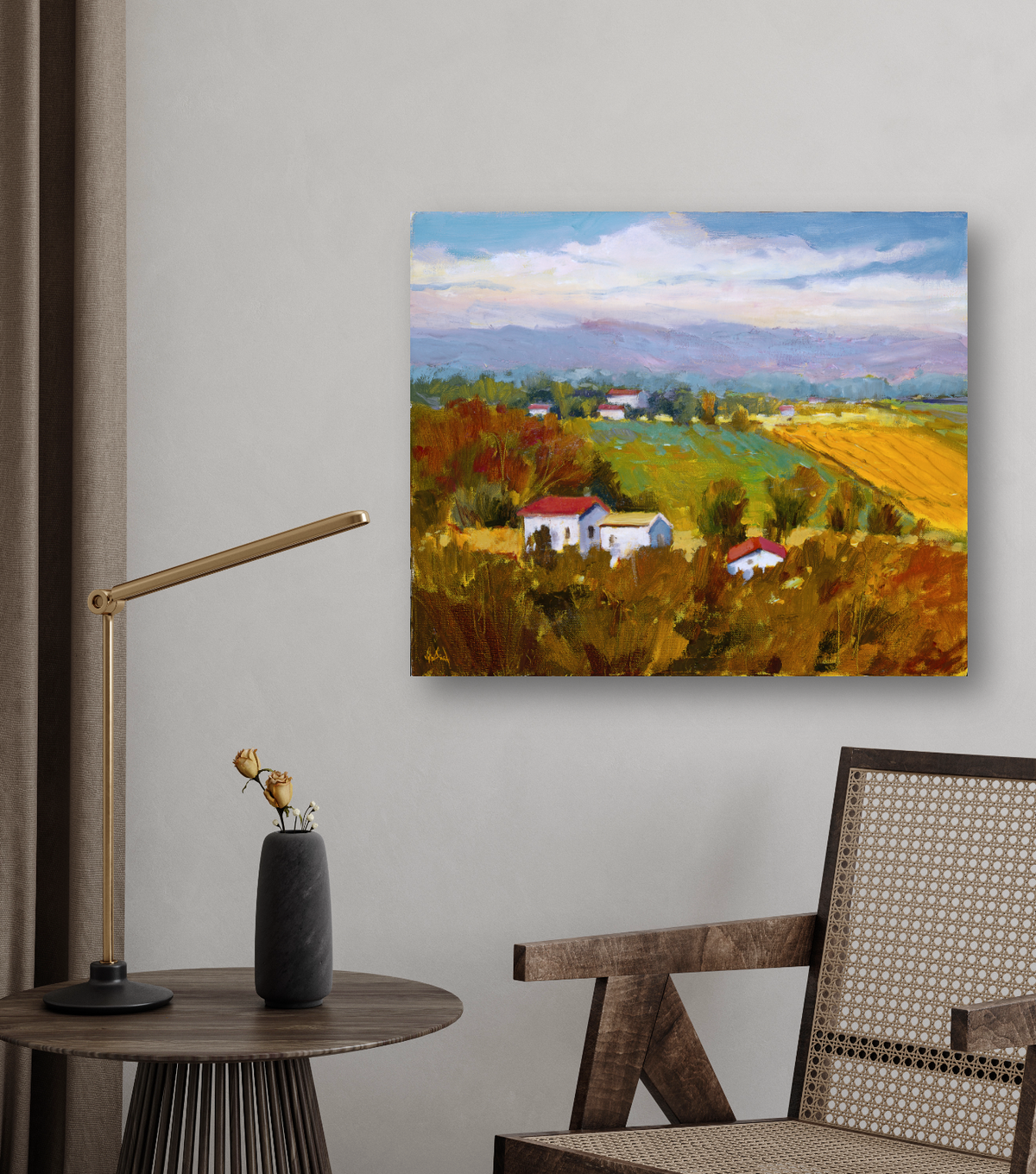 Canvas print of a vibrant rural landscape with green and golden fields, white houses with red roofs, and distant blue mountains under a cloudy sky, hung on a gray wall above a wooden chair and side table.