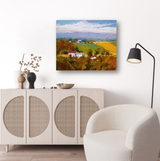 Canvas print of a rural landscape with green and golden fields, white houses with red roofs, under a partly cloudy blue sky, displayed above a light wood cabinet.