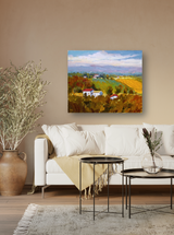 Colorful landscape painting of a plowed field with white houses, green trees, and a bright blue sky, hung on a beige wall above a white sofa.