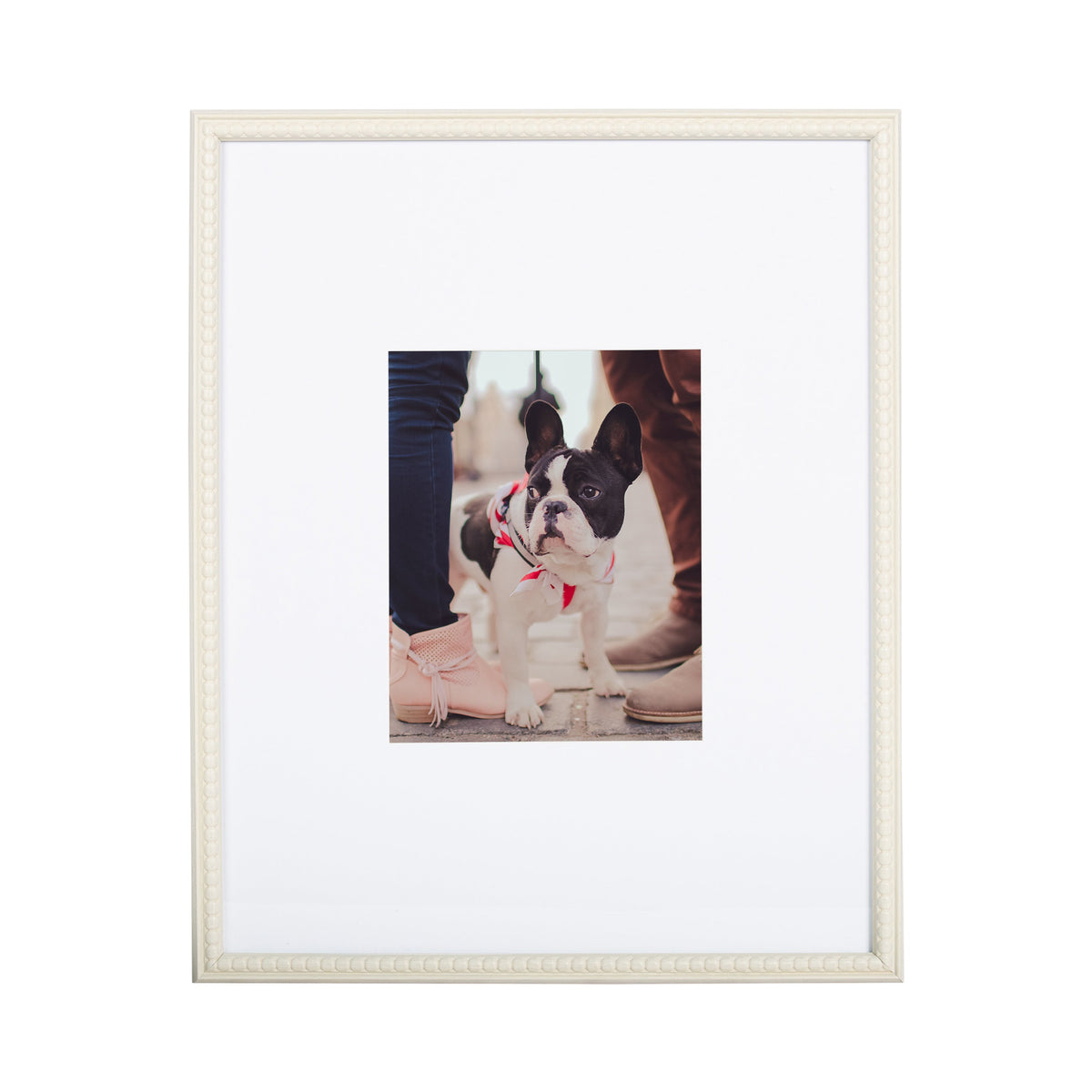 White photo frame with a beaded border enclosing a small photo of a black and white French bulldog wearing a red harness, between two pairs of legs with blush and beige shoes.