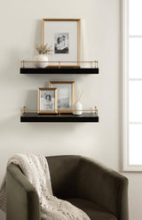 Two black floating shelves with thin gold railings on a white wall, holding framed black-and-white photos, a white vase with sticks, and a small white pot next to an olive green armchair with a knitted cream throw.
