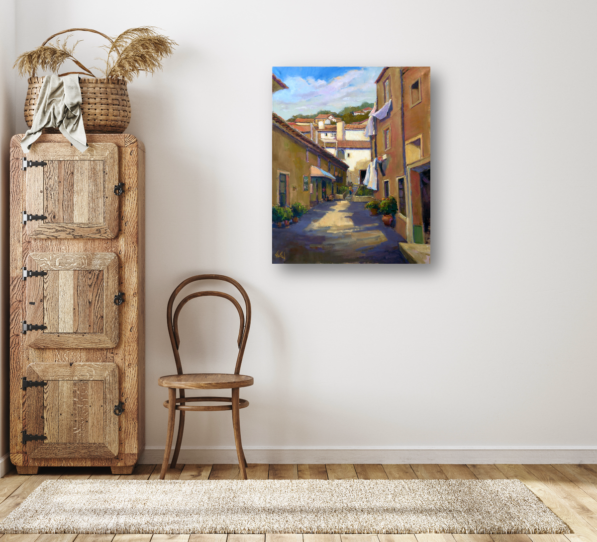Canvas print of a sunlit narrow alley with warm-toned buildings and greenery, hanging on a white wall above a wooden chair and beside a rustic cabinet.