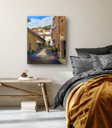 Canvas print of a sunlit narrow alleyway in a Mediterranean village with laundry hanging and potted plants along pastel-colored buildings, displayed above a wooden bench in a modern bedroom.