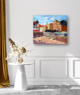 Canvas print of a vibrant Portuguese beach scene with golden sand, colorful buildings, palm trees, and a bright blue sky, displayed above a white wall with decorative molding.