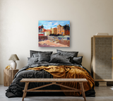 Bedroom with charcoal gray bedding and mustard throw, wooden bench and side table, rattan cabinet, and colorful painting of a coastal town on beige wall