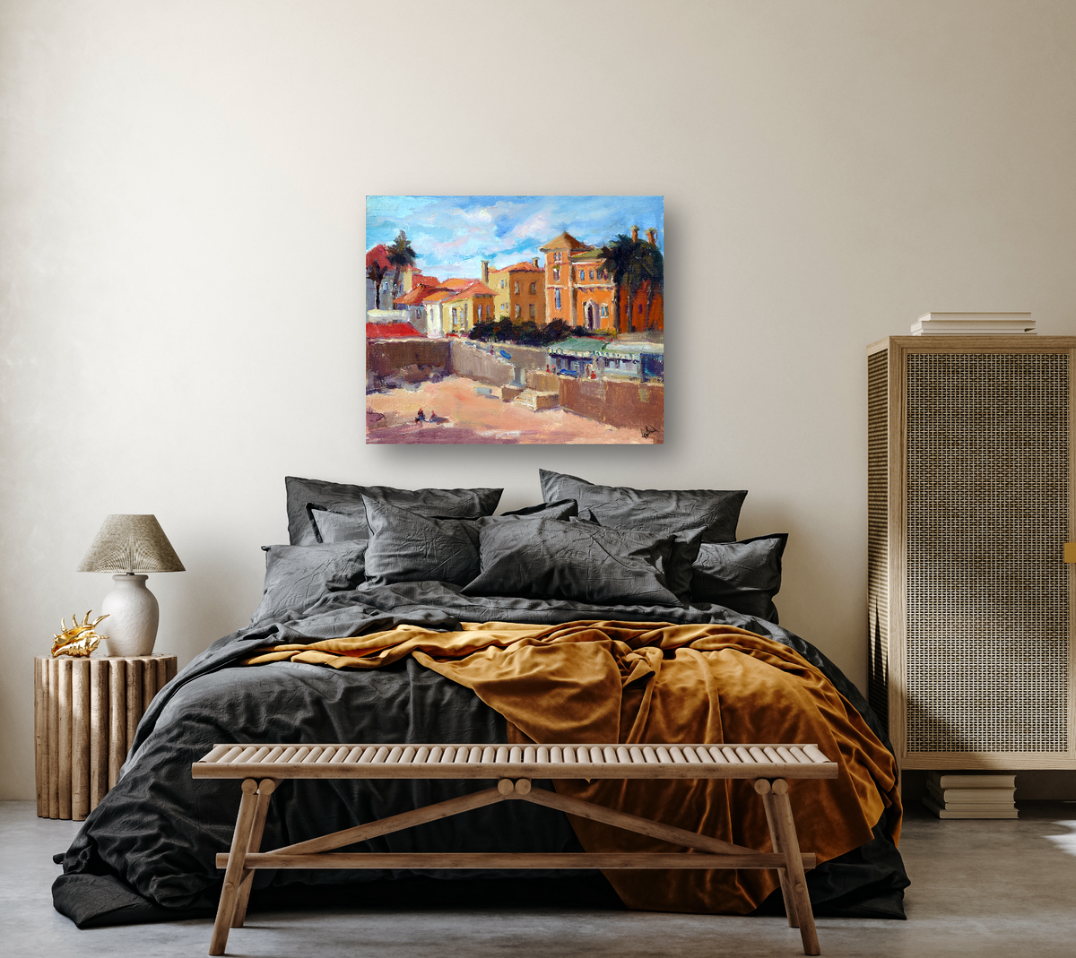Bedroom with charcoal gray bedding and mustard throw, wooden bench and side table, rattan cabinet, and colorful painting of a coastal town on beige wall