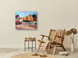Canvas painting of a Portuguese beach scene with buildings, stone stairs, and palm trees under a partly cloudy blue sky, displayed on a neutral wall above a woven armchair.