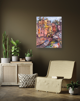 Colorful canvas print of a vibrant Italian coastal village with boats, displayed on a dark olive wall above a wicker chair and wooden cabinet with potted plants.