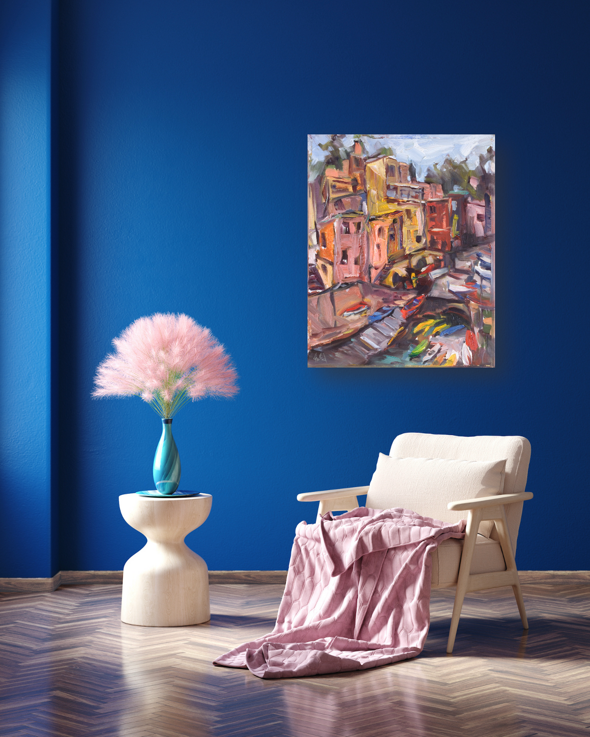 Abstract canvas painting of colorful buildings in earthy tones on a vivid blue wall above a beige armchair with a mauve throw and a small round white side table holding a blue vase with pink pampas grass.