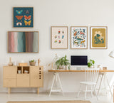 Home office wall with six framed artworks above light wooden desk and cabinet, featuring colorful butterflies, abstract colors, flowers, and animals in bright, muted tones.