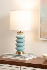 Table lamp with a stacked glossy light blue base and brushed gold accents under a plain white cylindrical shade, sitting on books on a wooden dresser.