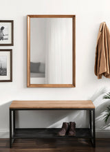 Rectangular wall mirror with a medium wood frame mounted above a bench combining matching wood top and black metal slatted shelf holding brown boots.
