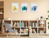 Three framed prints of patterned armchairs—yellow, teal, and blue—arranged horizontally above a white bookshelf filled with books and decor in a beige-walled room.