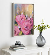 Framed canvas painting of vibrant pink flowers and tall purple blossoms against a soft, warm background, displayed on a white wall above a table with books and a gray vase.
