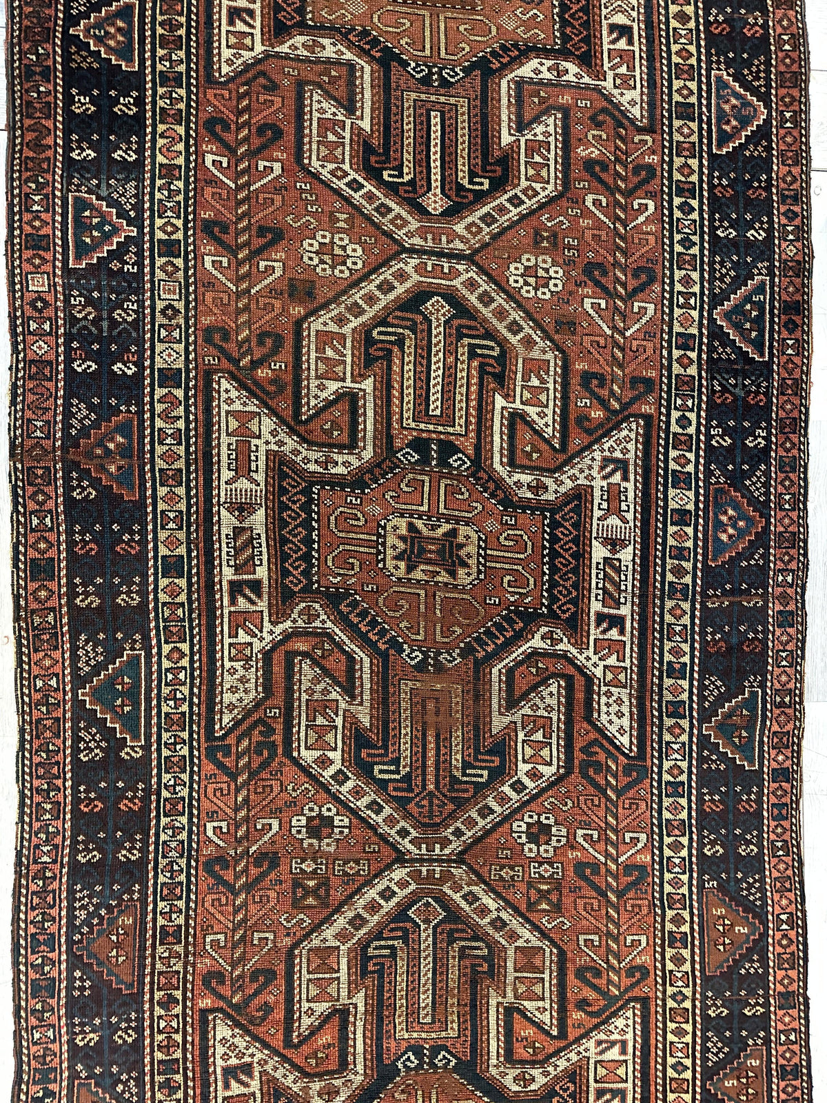 Narrow rug with three large geometric shield motifs in rust, cream, and dark blue, surrounded by multiple borders featuring small floral and diamond patterns.