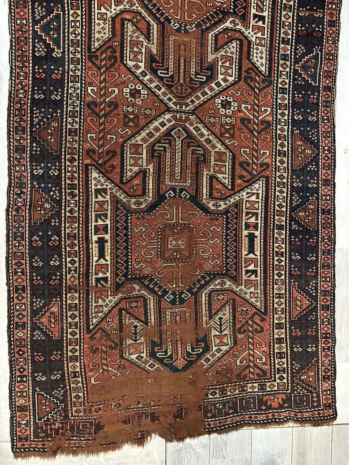 Antique Sewan Kazak rug with geometric triple shield motifs in rust, cream, and dark blue, bordered by repeating triangular patterns, showing wear at one end.