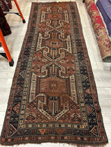 Long rectangular Sewan Kazak rug featuring repeating triple shield medallions in brown, black, and cream, with intricate geometric border patterns on a wooden floor.