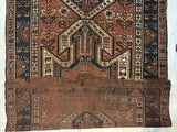 Pre-1900 Triple Shield Sewan Kazak rug with geometric motifs in rust, navy, and cream, featuring a central shield design and intricate border patterns, shown partly folded.