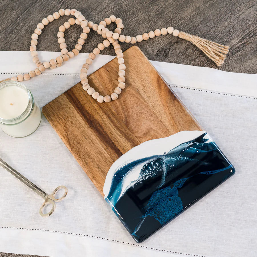 Wooden resin charcuterie board with blue resin design on a white surface with a candle and beads.